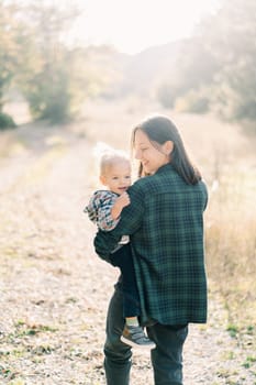 Smiling mother carries her little daughter in her arms while walking on the lawn and looks at her. Back view. High quality photo