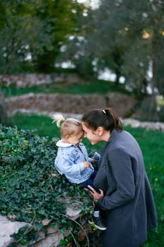 Mom and little girl touch their foreheads while sitting on a stone fence. High quality photo