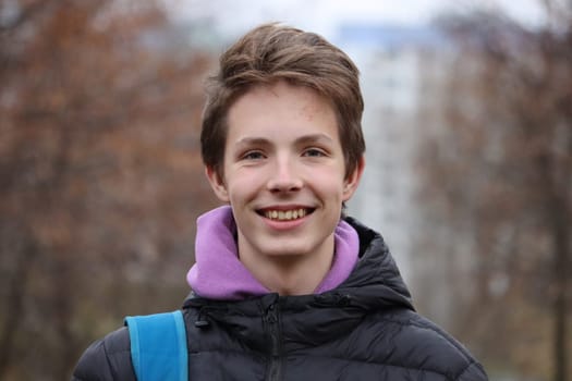 Close-up portrait of a cute and smiling boy 14-17 years old in a black jacket against the background of autumn trees without leaves, bokeh.