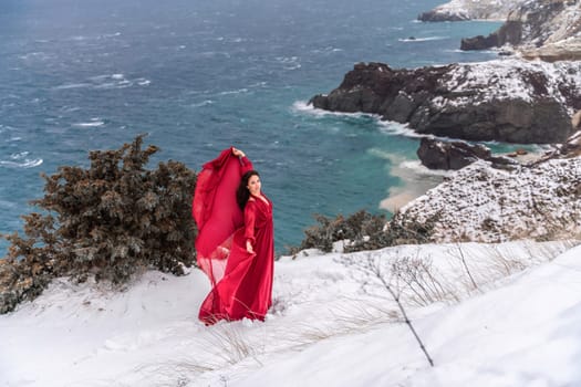 Woman red dress snow sea. Happy woman in a red dress in the snowy mountains by the emerald sea. The wind blows her clothes, posing against sea and snow background