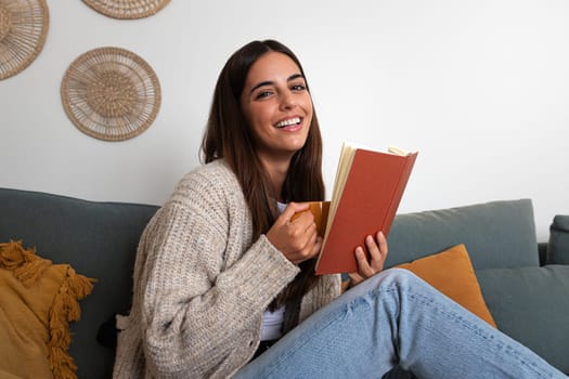 Relaxed woman reading a book at home, drinking coffee sitting on the couch looking at camera. Lifestyle concept.