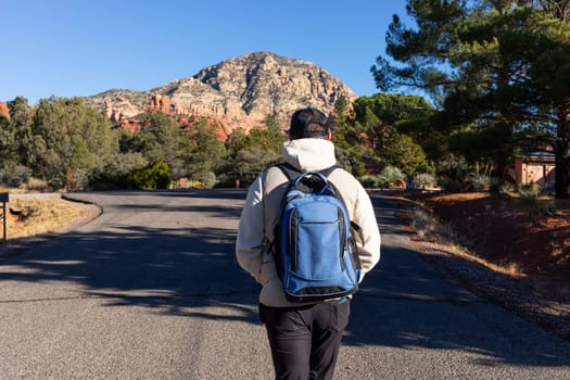 Back View of Male Tourist with Backpack Walking the Road Towards Red Rock Mountain Butte Of Sedona, Arizona, USA Beautiful Scenic Landscape. Tourism and Attraction, Spiritual Place. Horizontal Plane
