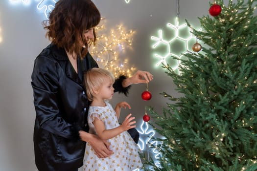 A mother with a 2-year-old daughter decorates the Christmas tree. Mom in a black suit, a girl in a white dress, her daughter hangs a red ball on the Christmas tree. Merry Christmas and New Year concept.