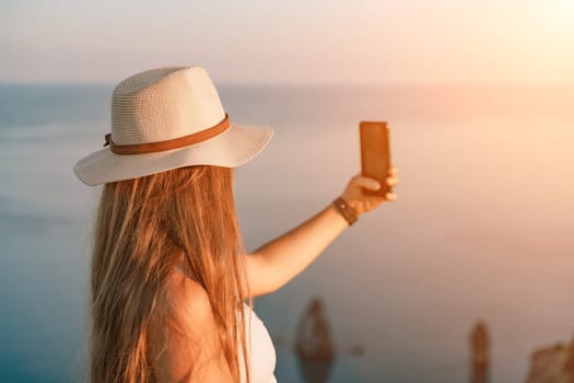 Selfie woman in a hat, white tank top, and shorts captures a selfie shot with her mobile phone against the backdrop of a serene beach and blue sea