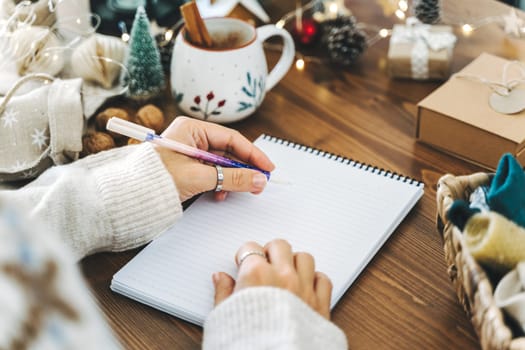 Woman hand holding pen on notepad at home on winter holidays xmas. Goals plans make to do and wish list for new year christmas concept, girl writing in notebook. Christmas decoration, gift boxes.
