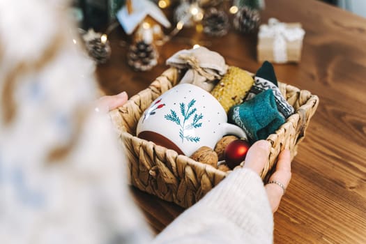 Woman s hands wrapping Christmas eco gift wicker basket, close up. Unprepared presents on wooden table with natural decor elements and items Christmas packing Concept.