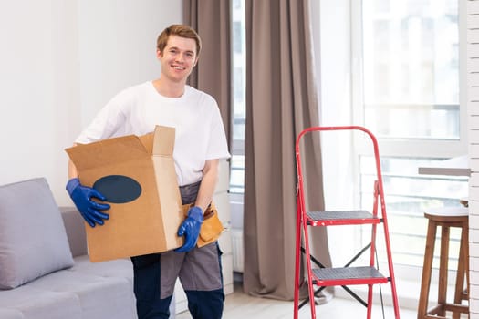 Smiling male worker of moving and delivery company holding cardboard box. Loader in overalls posing against background of colleague who packs cardboard boxes. Moving service concept