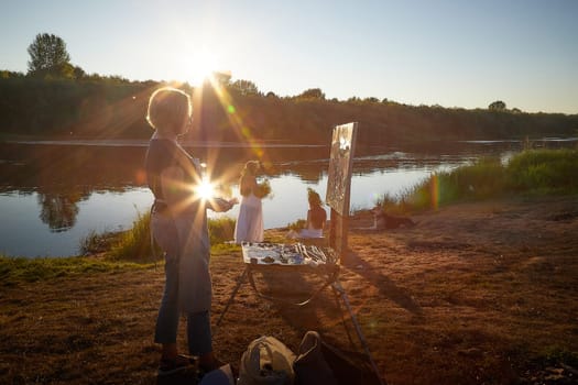 Adult female artist painting picture near water of river or lake in nature and girls in white sundress and flower wreath. Artist and models posing in holiday of Ivan Kupala in nature at sunset