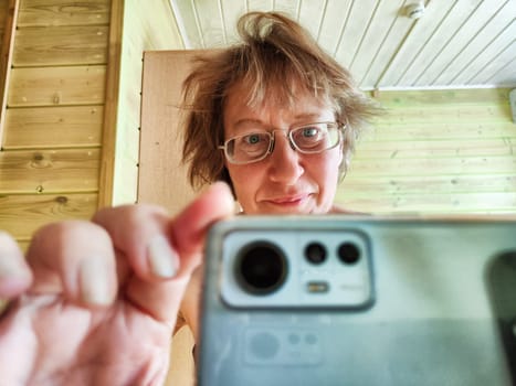 A shaggy, funny middle-aged woman in glasses takes selfie on her smartphone in front of the mirror in the morning. A blogger girl with disheveled hair takes pictures of herself for a blog