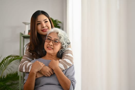 Beautiful moment of affectionate young woman hugging senior mother from back. International hug day concept.