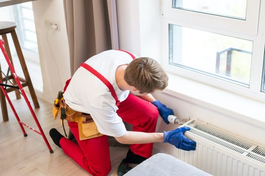 Young handsome professional plumber worker installing heating radiator in an empty room of a newly built apartment or house. Construction, maintenance and repair concept.