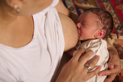 Close-up of a young woman breastfeeding her newborn baby at home. Selective focus on the baby sucking mother's breast. Parenthood, motherhood, maternity leave.