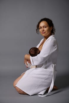Portrait of a confident authentic happy mother holding her newborn baby, breastfeeding him, smiling cutely looking confidently at the camera, sitting isolated over gray studio background