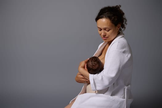 Happy woman in white waffle bathrobe, holding and feeding her newborn baby, sitting isolated over gray studio background with copy ad space. The mother breast milk is the best nourishment for baby