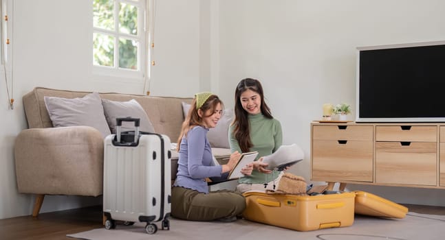 Two young Asian women are prepare for the journey happily. Pretty girls are packing suitcase for the trip and check the list of various items.