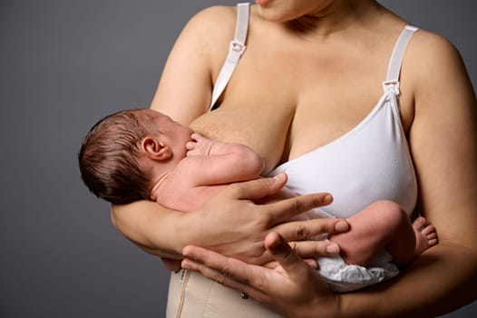 Selective focus on a 2 week old adorable newborn baby suckling at his mother's breast, isolated over gray studio background. Breastfeeding and healthy lactation concept