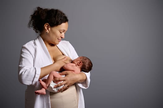 Authentic portrait of a curly haired woman in white bathrobe and bandage after cesarean, holding her newborn baby suckling at her breast, feeding him with mother milk, isolated gray studio background