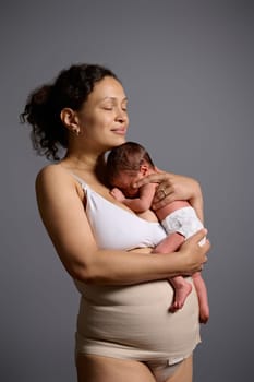 Delightful mixed race woman, happy loving caring mother hugging her newborn baby, standing together isolated gray studio background. Young mom in elastic bandage after cesarean section with her child