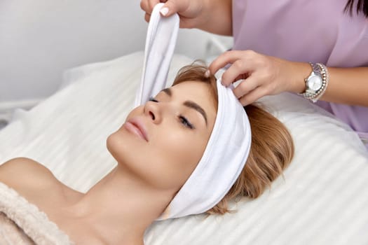 Cosmetologist putting white headband towel on female patient head in beauty salon