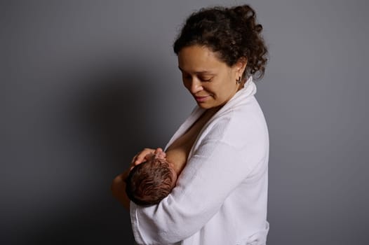 Authentic studio portrait of African American pretty woman, young mother smiling looking at her newborn baby, while breastfeeding him. People. Maternity leave. Healthy lifestyle