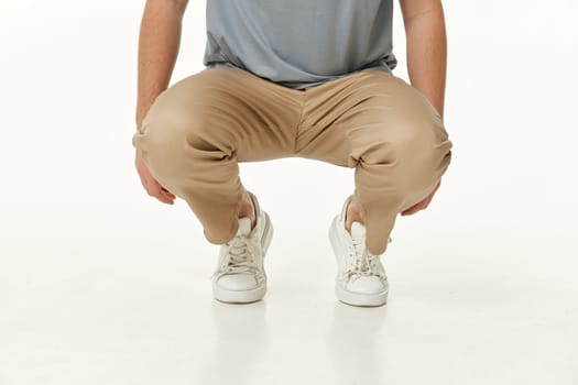 man wearing white sneakers and casual beige pants squatting on studio background. front view