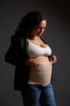 Charming multi ethnic young woman wearing an elastic bandage on her postnatal belly, isolated gray background. Copy space. Postpartum recovery after cesarean c-section. Maternity lifestyle. Body care