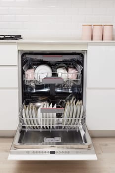 Open built-in dishwasher machine with clean cutlery, dishes, plates in white modern kitchen. front view