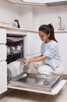 pretty woman loading dished to open automatic built-in dishwasher machine with clean utensils inside in modern white kitchen.