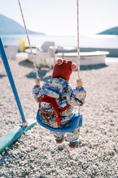 Small child in overalls with a backpack swings on a swing on a sunny beach and looks at the sea. High quality photo