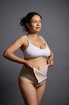 Authentic studio portrait of a multi ethnic woman, young mother putting on an elastic bandage after cesarean c-section, looking away, isolated over gray studio background. Skin and body care concept