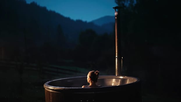 Woman bathes alone in open-air bath with night mountains view. Wooden hot tub outside. Lady enjoying recreation, spa procedures in steaming pool, private treatment.