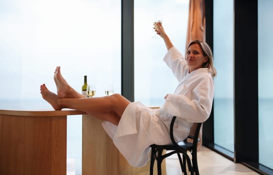 Woman in bathrobe with feet on table drinking wine from glass at home. Rest and relaxation with alcohol after spa treatments concept