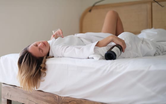 Drunk woman in bath robe lying in bed with bottle of wine in hotel. Loneliness and alcohol addiction in women concept