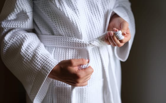 Women hands tying belt on white bathrobe closeup. Comfortable clothes for home concept