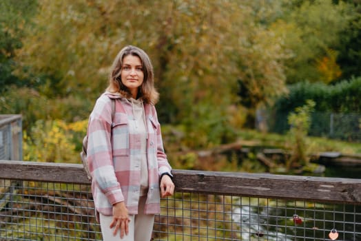 Portrait of cute young woman in casual wear in autumn, standing on bridge against background of an autumn Park and river. Pretty female walking in Park in golden fall. Copy space. smiling girl in the park standing on wooden bridge and looking at the camera in autumn season