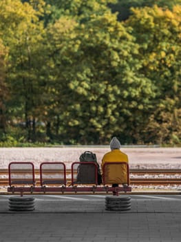 Man with travel backpack sits on bench at station waiting for train. Village train station against thick autumn forest with passenger waiting for train
