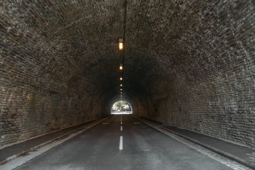 Infrastructure demonstrates auto tunnel illuminated by lights hanging on ceiling. City road inside stone mountain with light exit at end