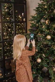 A little blonde girl photographs balloons on a Christmas tree in a festive interior decorated in a New Year's style. The concept of a merry Christmas