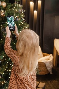 A little blonde girl photographs balloons on a Christmas tree in a festive interior decorated in a New Year's style. The concept of a merry Christmas