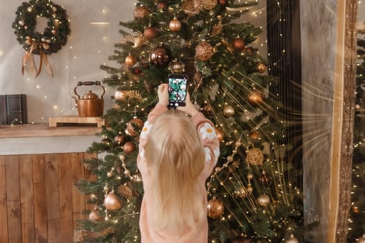 A little blonde girl photographs balloons on a Christmas tree in a festive interior decorated in a New Year's style. The concept of a merry Christmas