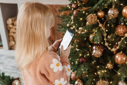 A little blonde girl photographs balloons on a Christmas tree in a festive interior decorated in a New Year's style. The concept of a merry Christmas