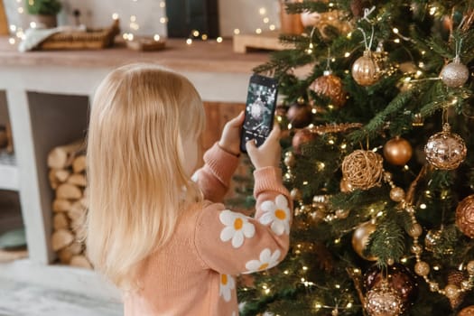 A little blonde girl photographs balloons on a Christmas tree in a festive interior decorated in a New Year's style. The concept of a merry Christmas