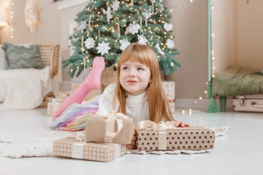 A little red-haired girl lies next to Christmas gifts in a craft package. Happy Christmas concept