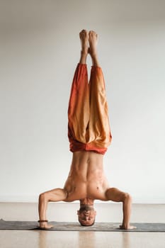 a man with a naked torso does yoga standing on his head indoors. Fitness Trainer.