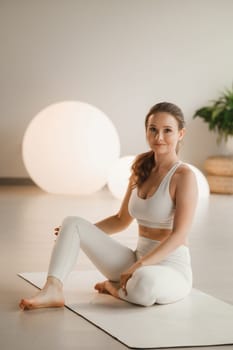 Portrait of a girl in white clothes sitting on a mat before doing Yoga indoors.