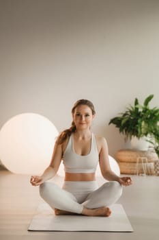 Portrait of a girl in white clothes sitting in a lotus position on a mat at an indoor yoga class.