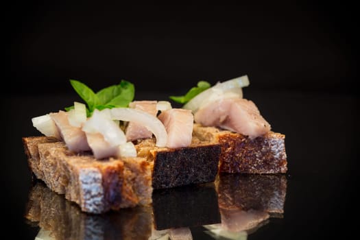 canape of pieces of salted herring with onions on a fresh dark piece of bread, isolated on a black background.