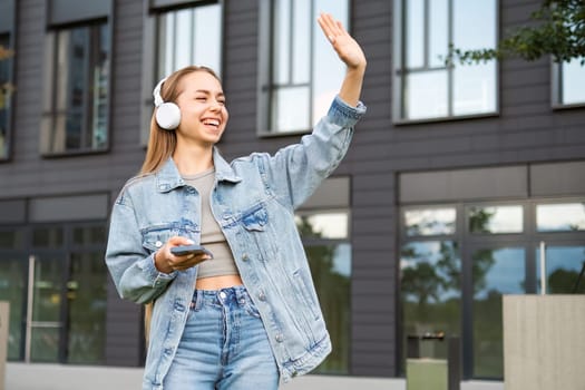 Young woman with a smile and headphones waves to someone on the street.