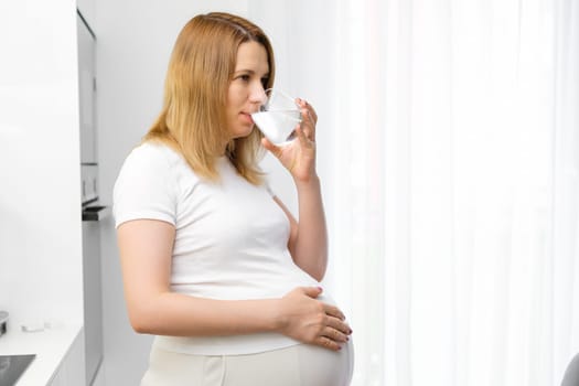 Good looking pregnant woman drinks water standing on the kitchen.