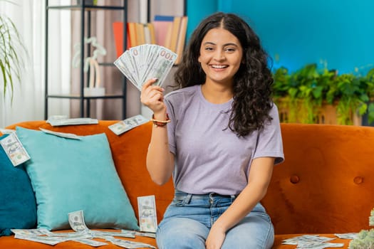 Successful happy rich Indian Arabian woman counting money cheering up with high profits, lottery game win, satisfied of wealth income earnings salary on home couch full of dollar cash banknotes